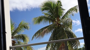 A balcony with two wicker chairs and a round table, overlooking palm trees and a clear blue sky.