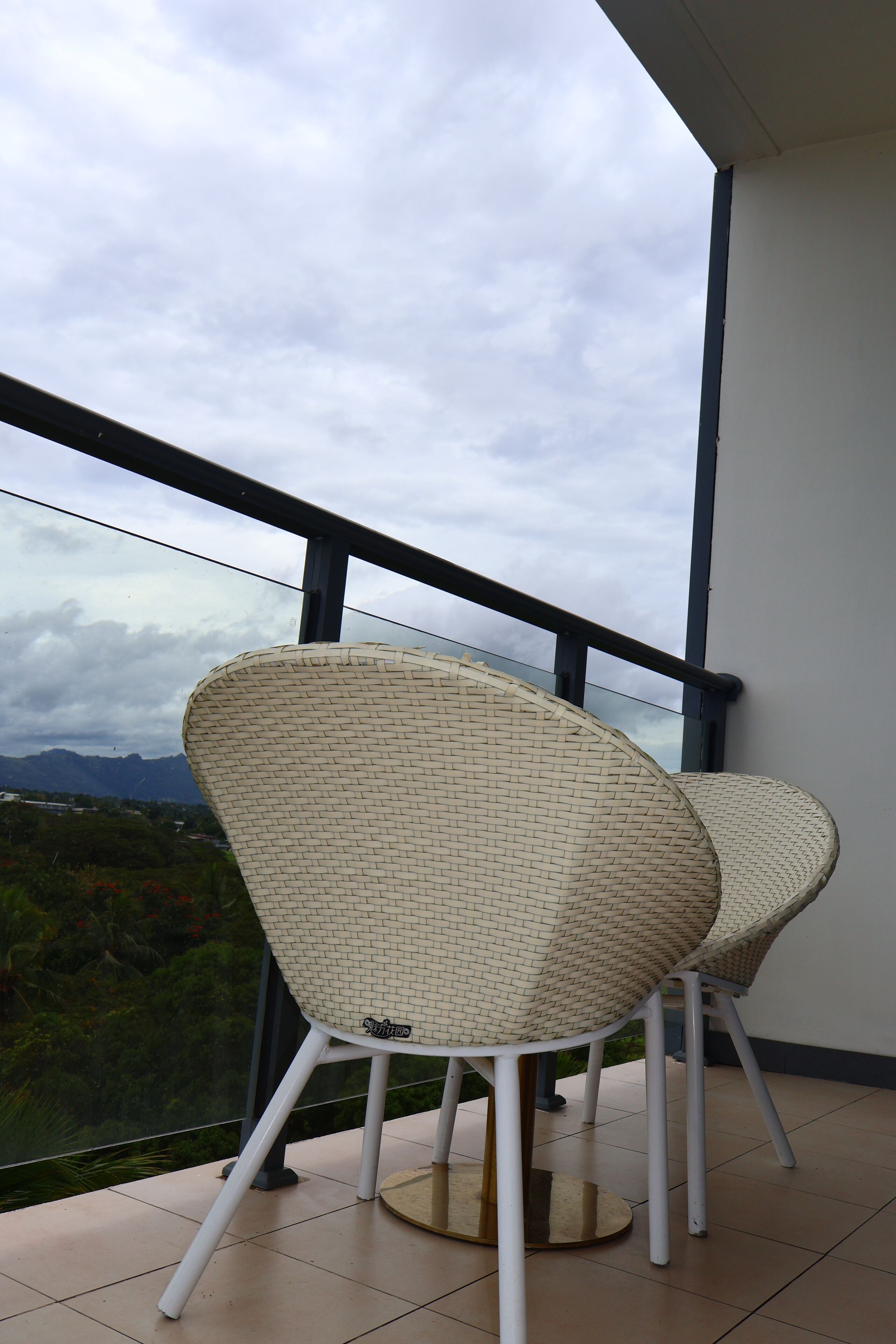 Two wicker chairs with metal legs on a balcony with a glass railing, overlooking a mountainous landscape.