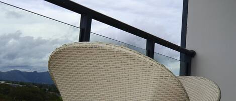 Two wicker chairs with metal legs on a balcony with a glass railing, overlooking a mountainous landscape.