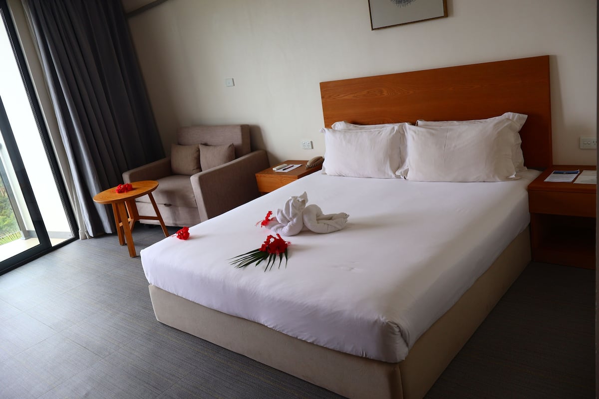 Une chambre d’hôtel avec un grand lit, une tête de lit en bois, un coin salon avec un fauteuil et une petite table, et une fenêtre avec des rideaux.