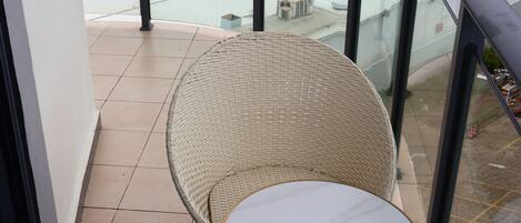 A balcony with a wicker chair and table, overlooking a building with a flat roof and a view of a green landscape.