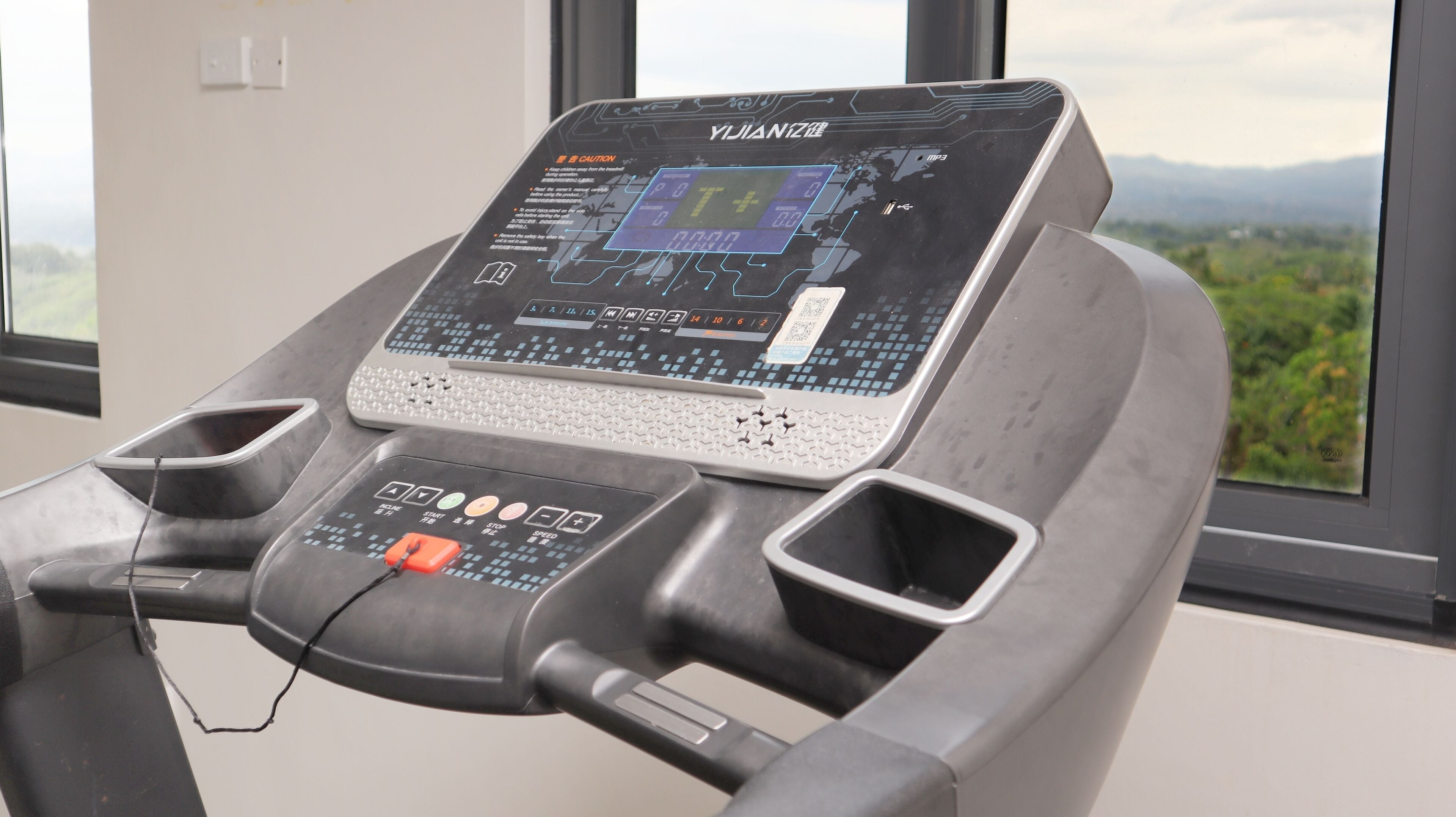 A treadmill with a digital display and control panel, positioned near a window with a view of greenery.