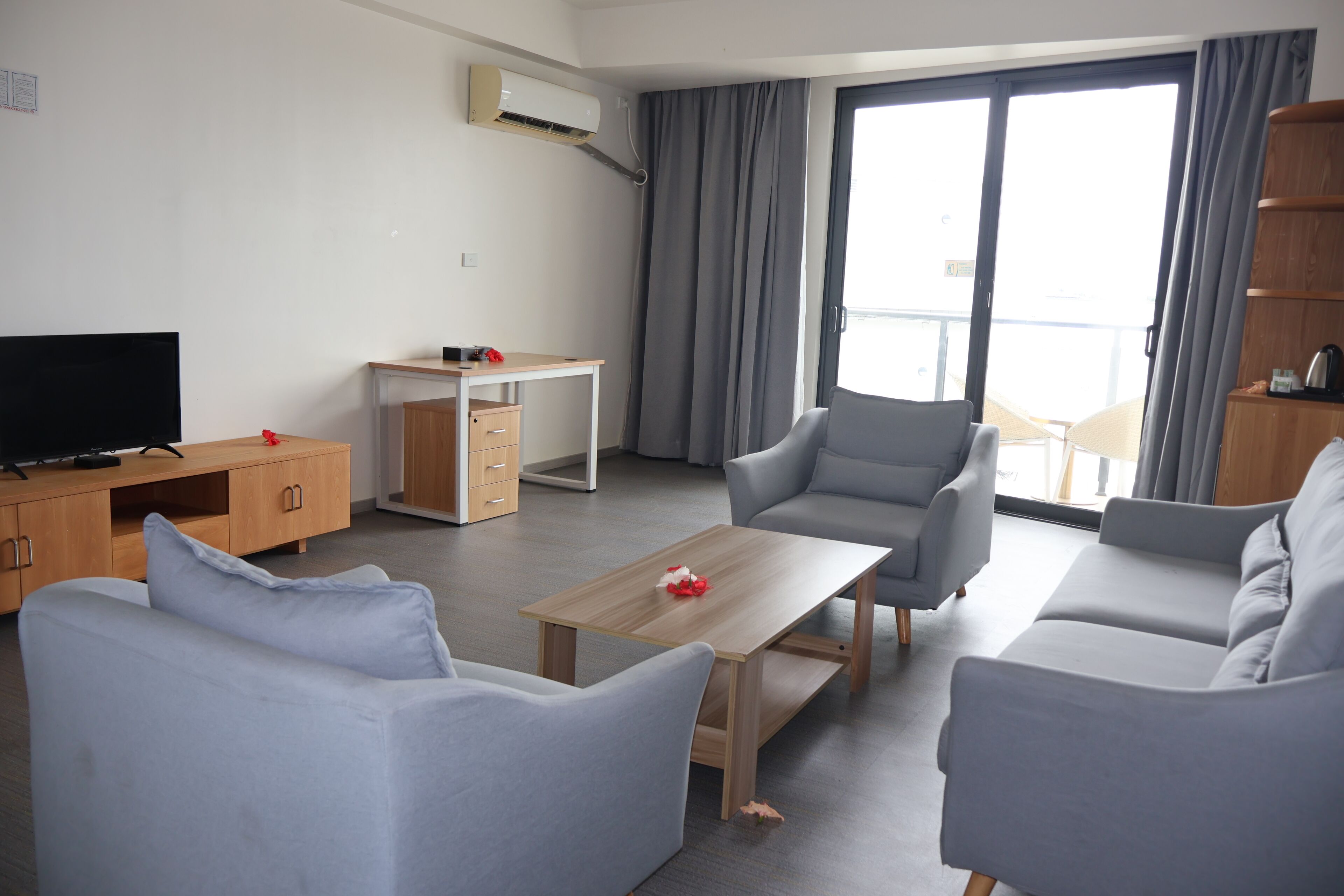 A modern living room with a sofa, armchairs, a coffee table, a TV, a desk, and a balcony with curtains.