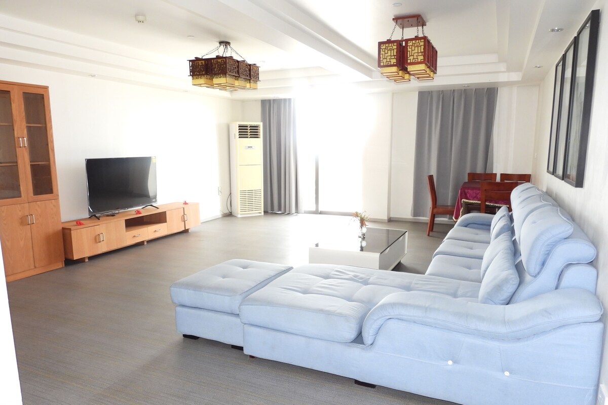 A modern living room with a sectional sofa, a flat-screen TV, wooden cabinets, and a dining area with a table and chairs.