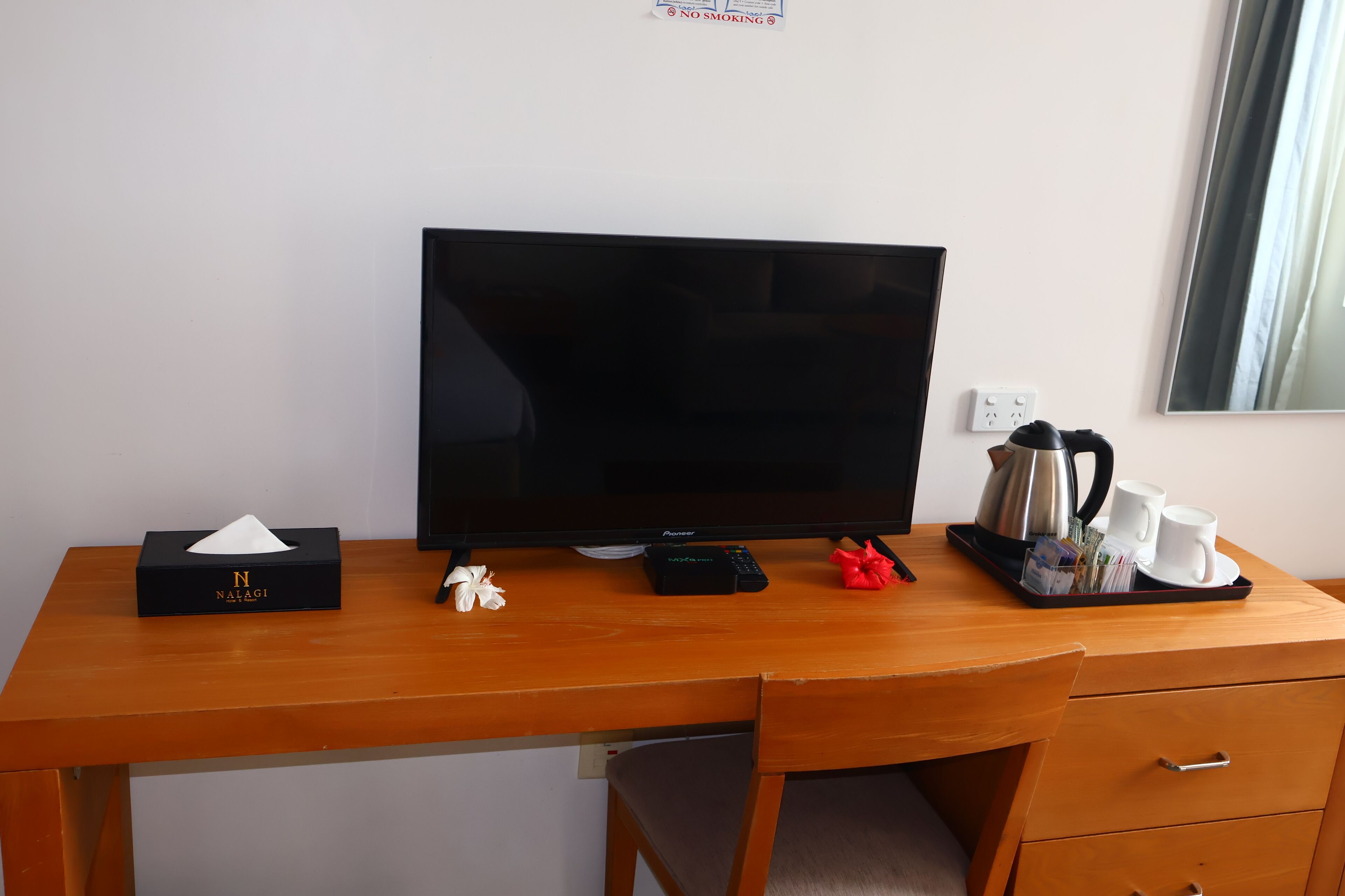 Habitación de hotel con un escritorio de madera, televisor, caja de pañuelos, control remoto, cafetera y una bandeja con tazas y un pequeño regalo rojo.