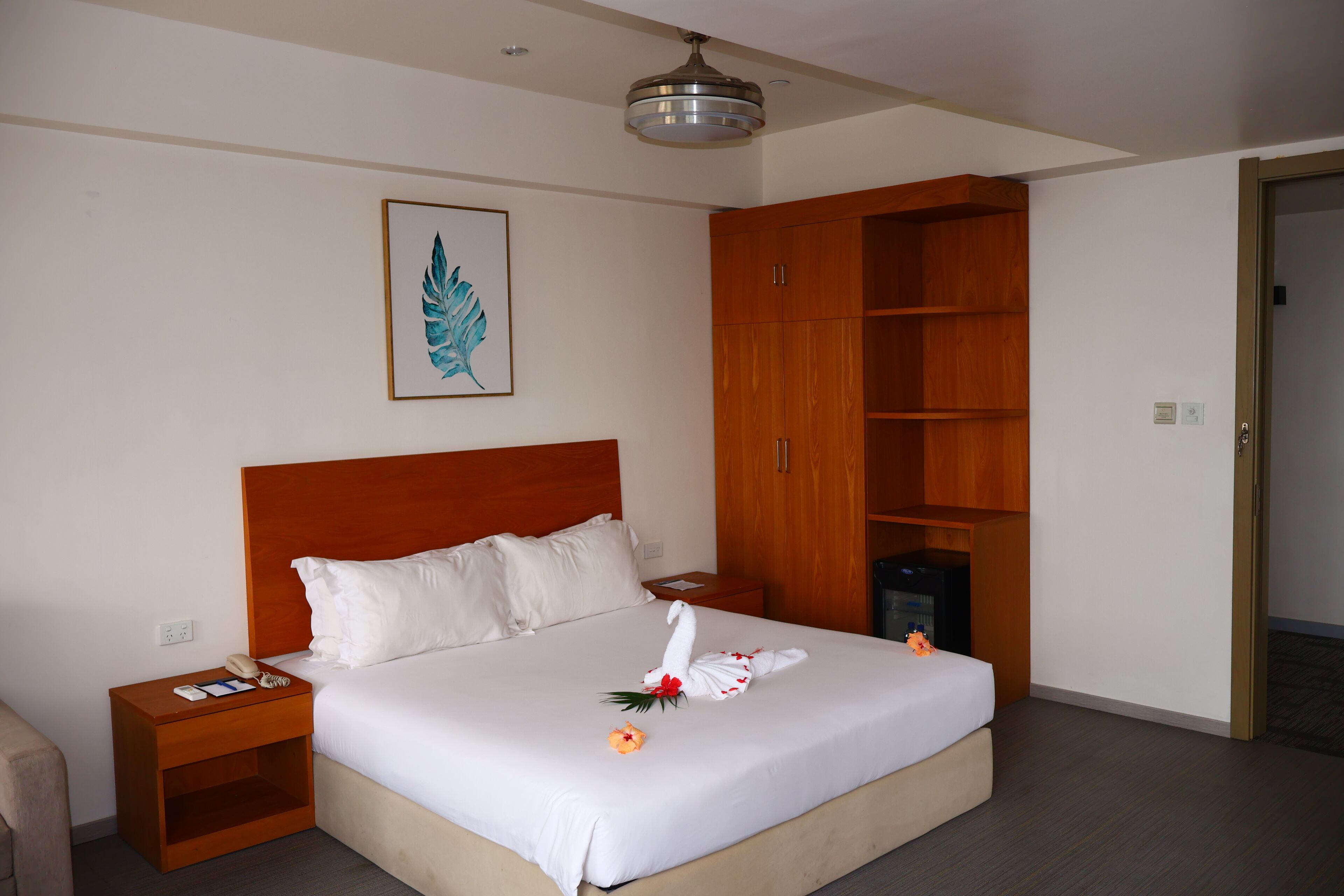 A hotel room with a large bed, wooden headboard, a nightstand with a phone, a framed artwork on the wall, and a wooden shelving unit.