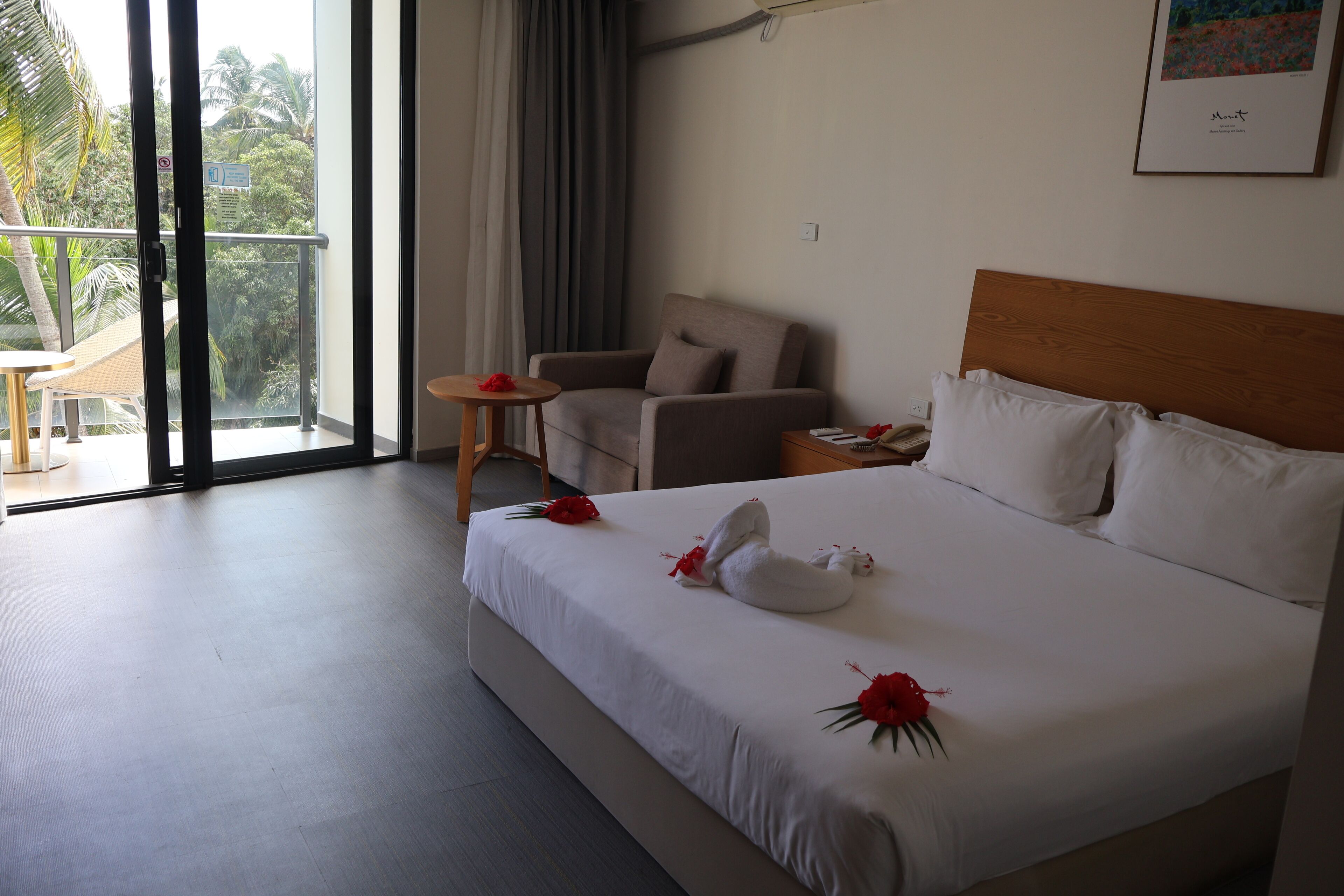 A hotel room with a bed, a chair, a small table, and a balcony with a view of palm trees.
