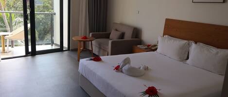 A hotel room with a bed, a chair, a small table, and a balcony with a view of palm trees.