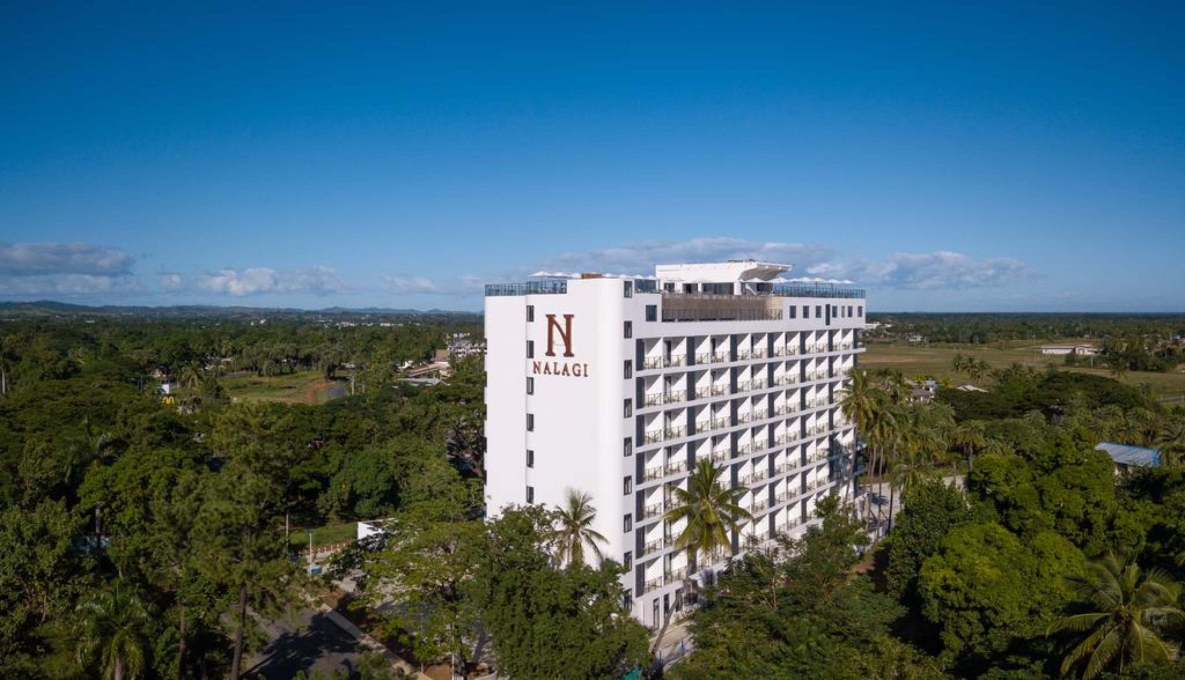 A high-rise hotel with a prominent 'N' logo on the side, surrounded by lush greenery and a clear blue sky.