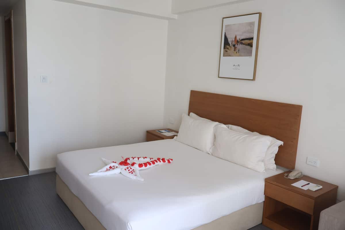 Une chambre d’hôtel avec un grand lit, une tête de lit en bois, une table de chevet avec un téléphone et des documents, et un tableau encadré au mur.
