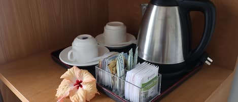 Una mesa de madera con un hervidor de acero inoxidable, tazas y platillos blancos, una flor de hibisco y una bandeja con bolsitas de té.