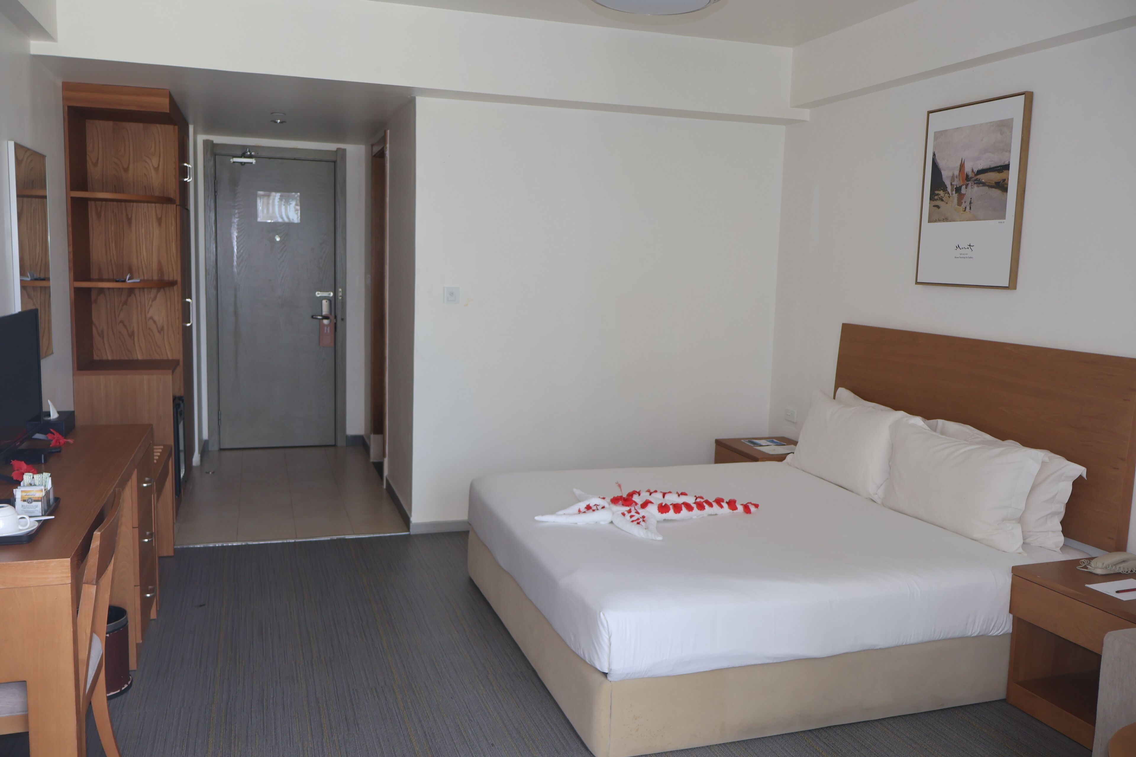 A hotel room with a bed, a wooden desk, a chair, a nightstand, and a framed picture on the wall.