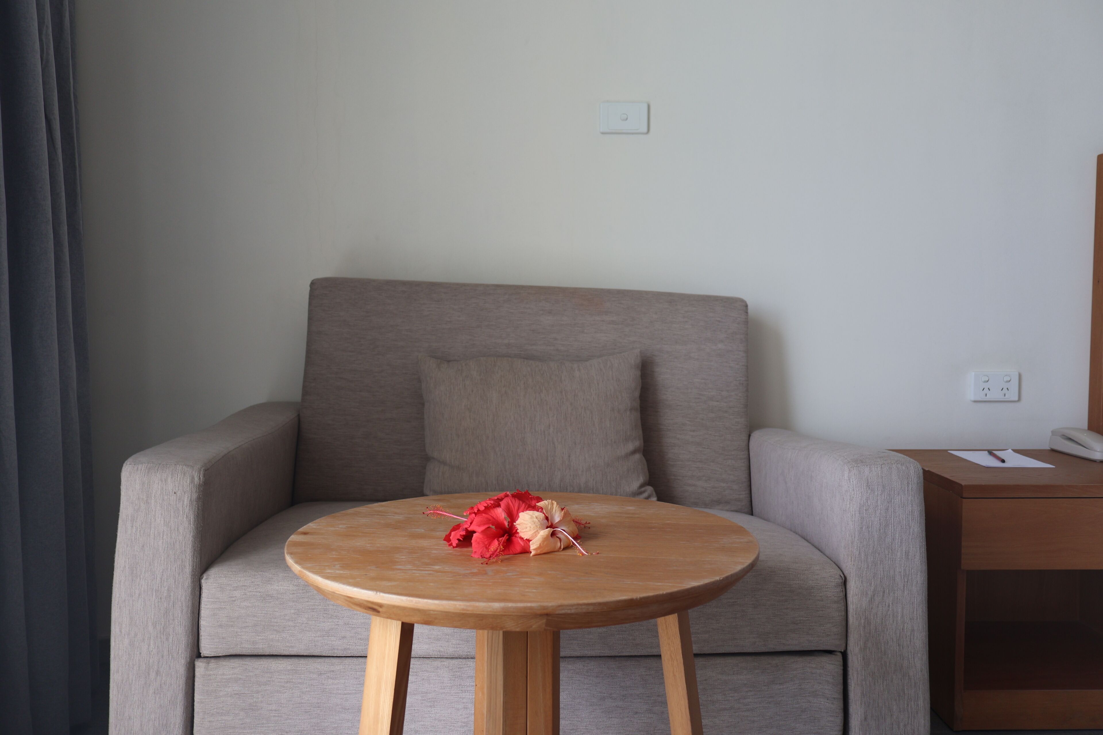 A modern room with a grey sofa, a wooden side table, and a round wooden table with a bouquet of red flowers.