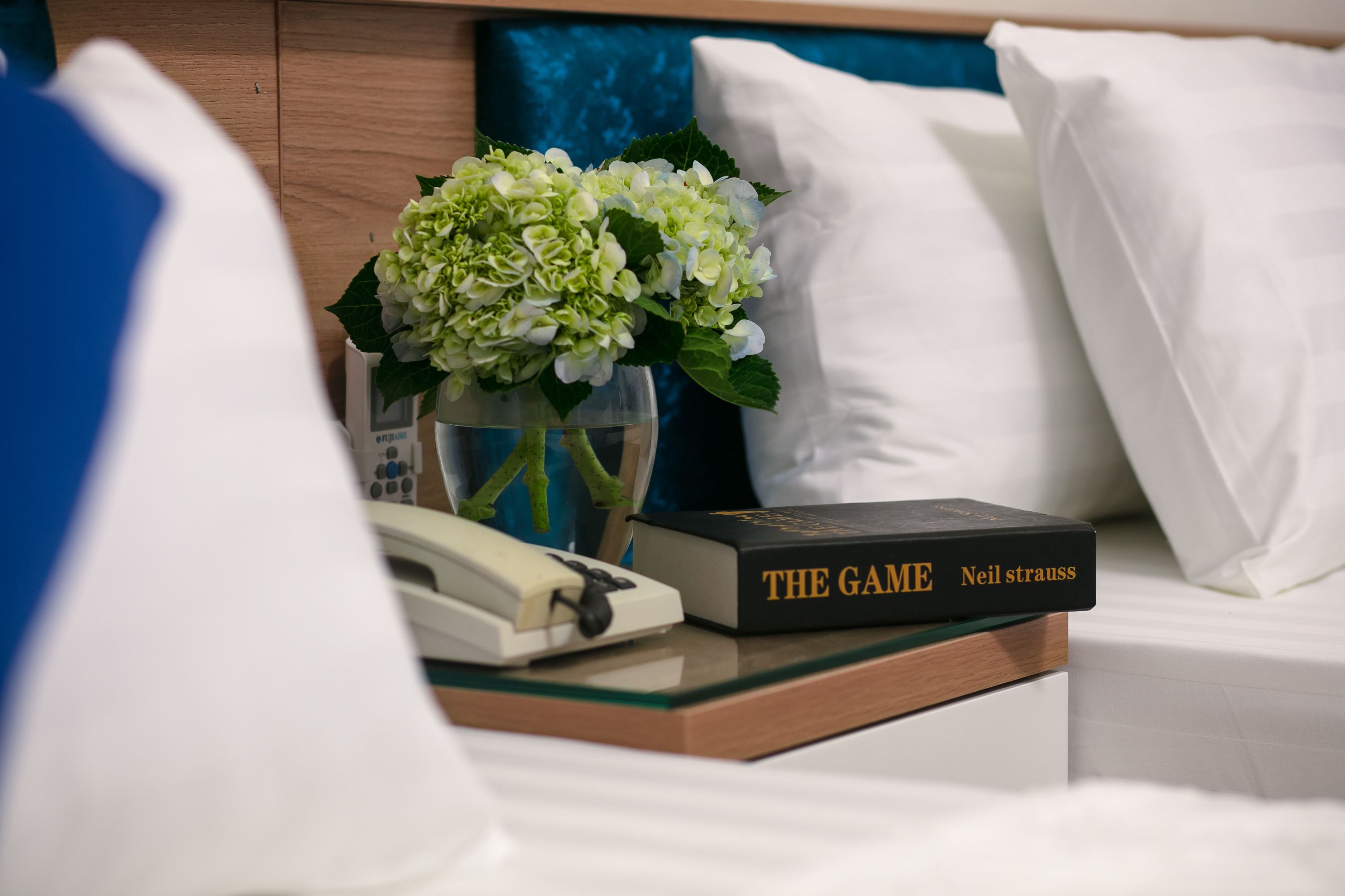 Habitación de hotel con una mesita de noche que sostiene un libro titulado 'The Game' de Neil Strauss, un florero con flores blancas y un teléfono.