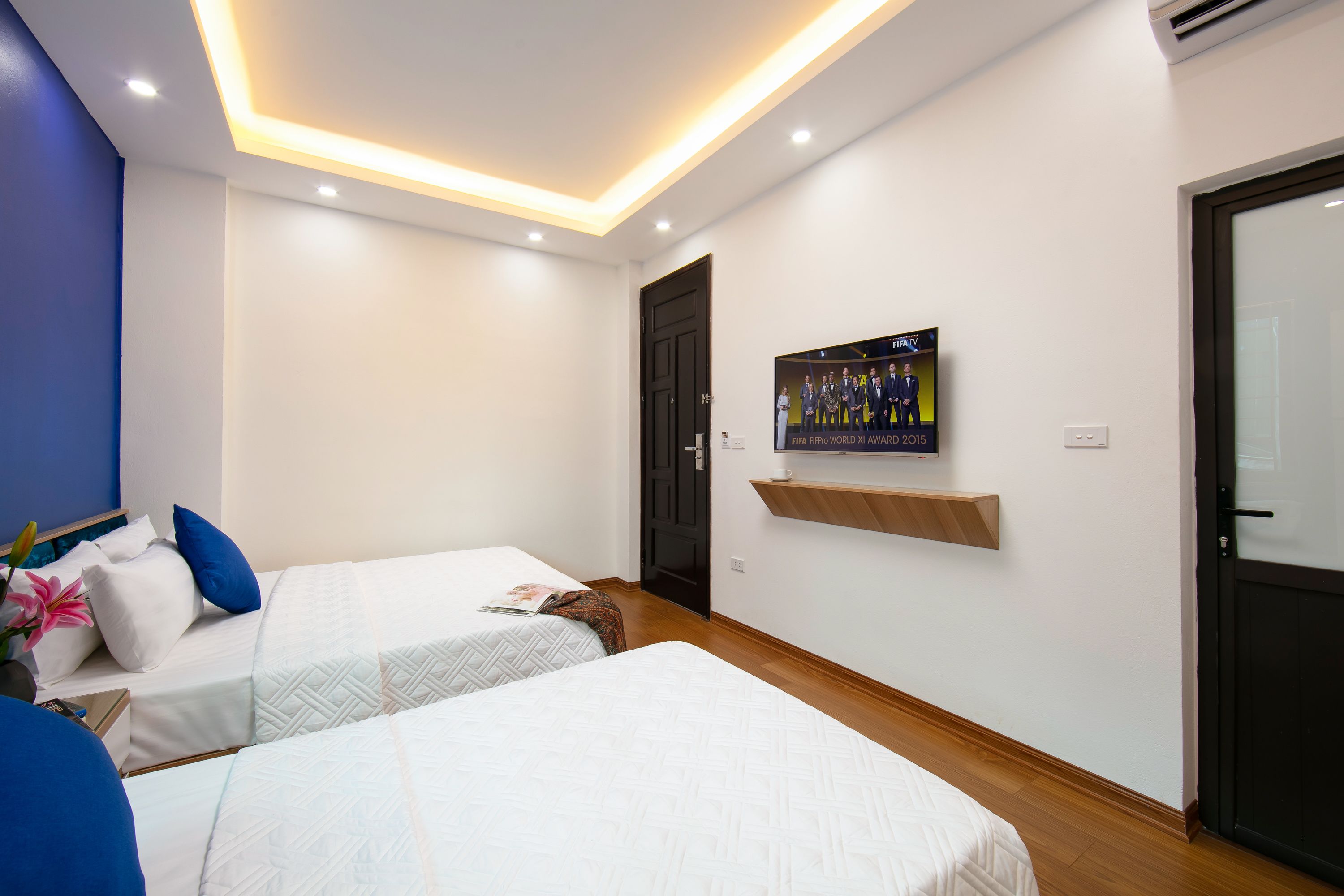 A hotel room with two beds, a TV mounted on the wall, a door, and a shelf with a FIFA poster.