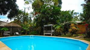 Ein Swimmingpool, umgeben von üppiger tropischer Vegetation, einer Holzterrasse und einem strohgedeckten Gebäude.
