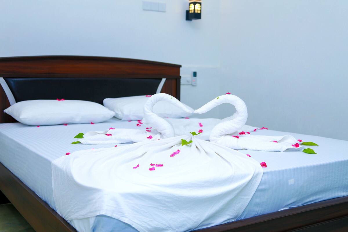 Una cama bien hecha con sábanas blancas y pétalos de flores rosa, un cabecero de madera y una lámpara fijada a la pared.