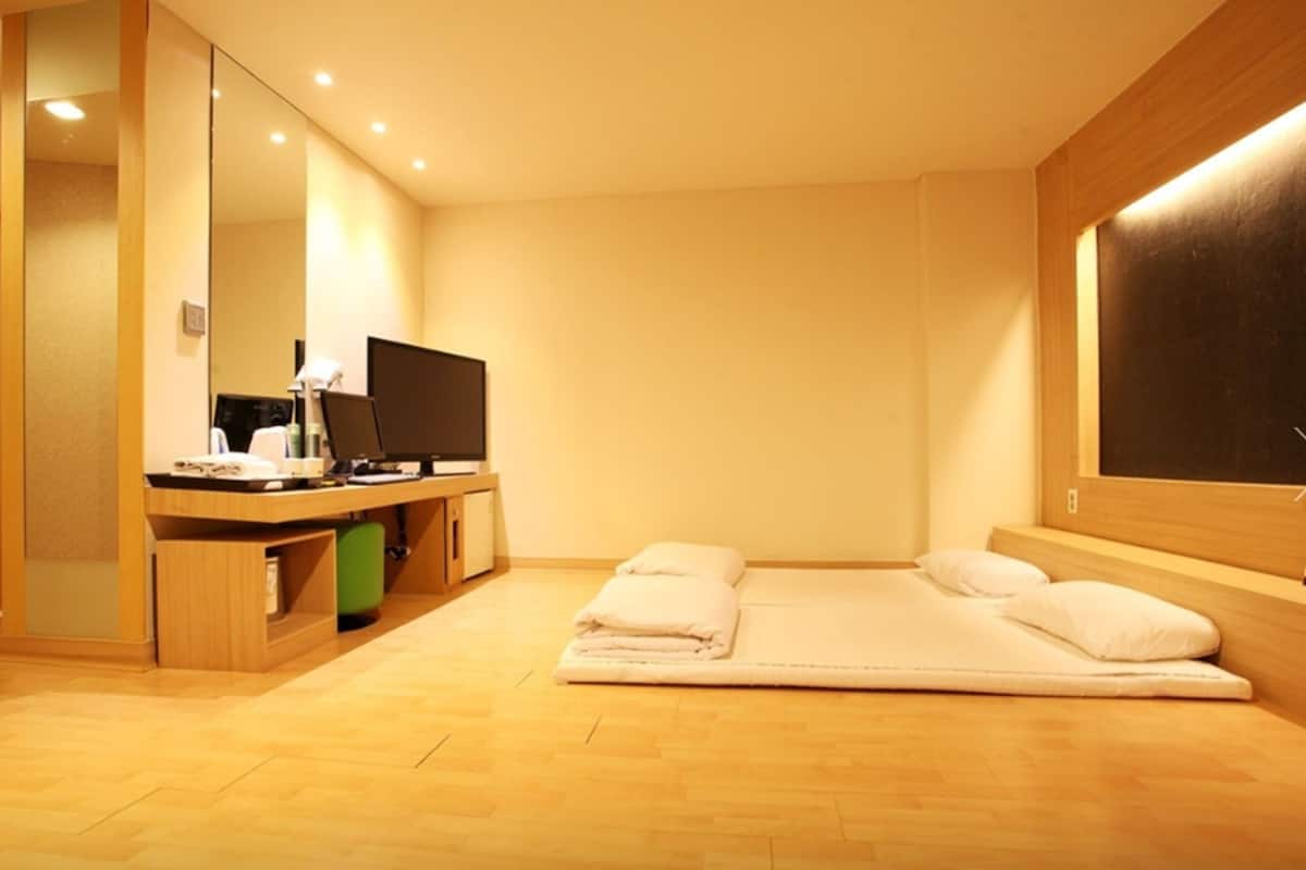 A hotel room with a wooden floor, a bed with white linens, a desk with a computer and a flat-screen TV, and a large mirror.