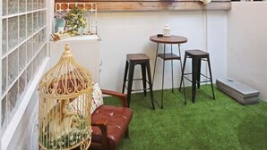 Un espacio al aire libre acogedor con pared de madera, una mesa pequeña, dos taburetes y una jaula de pájaros.