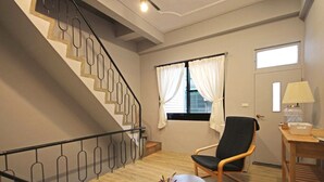 Una habitación con piso de madera, una silla negra, una mesa de madera con una lámpara, una ventana con cortinas blancas y una escalera con barandilla de metal.