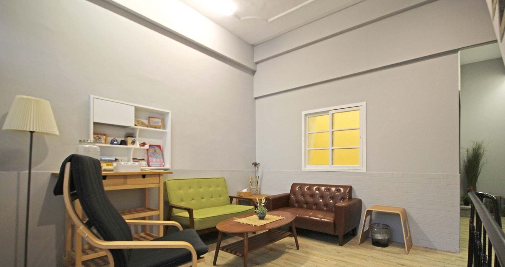 A living room with a green sofa, a brown leather sofa, a wooden chair, a small table, a lamp, and a window with yellow blinds.