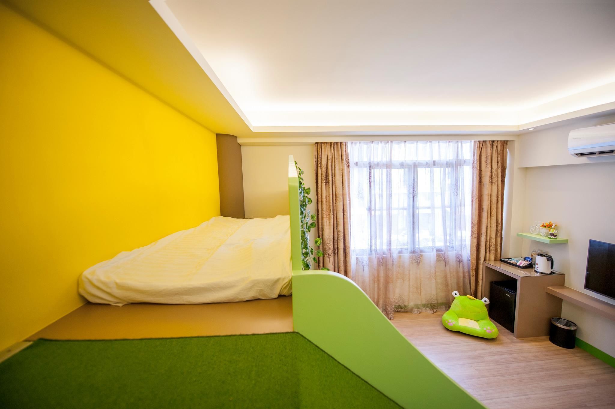 A compact hotel room with a bed, a small desk, a green bean bag chair, a TV, and a window with curtains.