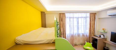 A compact hotel room with a bed, a small desk, a green bean bag chair, a TV, and a window with curtains.
