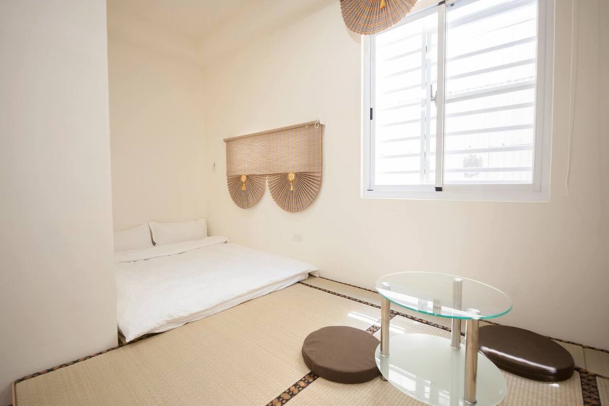 Ein schlicht eingerichtetes Zimmer mit einem Bett, einem runden Glastisch und einem Fenster mit Jalousien.
