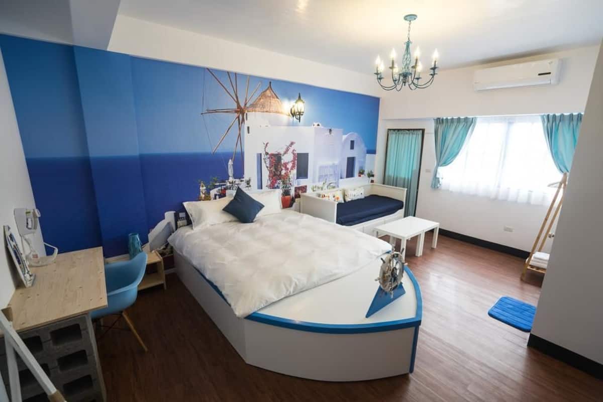 A modern bedroom with a curved bed, a desk, and a chair. There is a large window with curtains, a chandelier, and a wall mural.