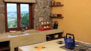 A kitchen with a stone wall, a window with a view, a blue kettle, and a shelf with kitchen utensils. - La Hacienda Old Grand Port Mauritius (Vieux Grand Port)