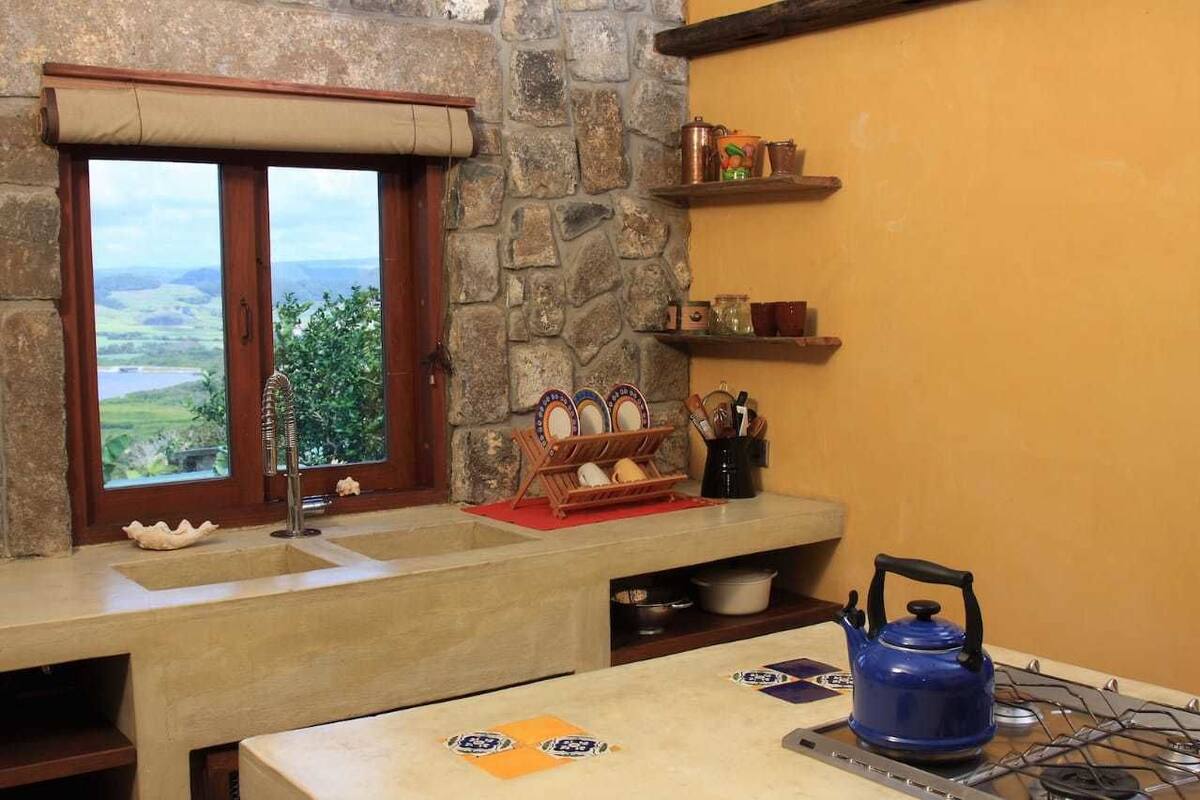 A kitchen with a stone wall, a window with a view, a blue kettle, and a shelf with kitchen utensils.