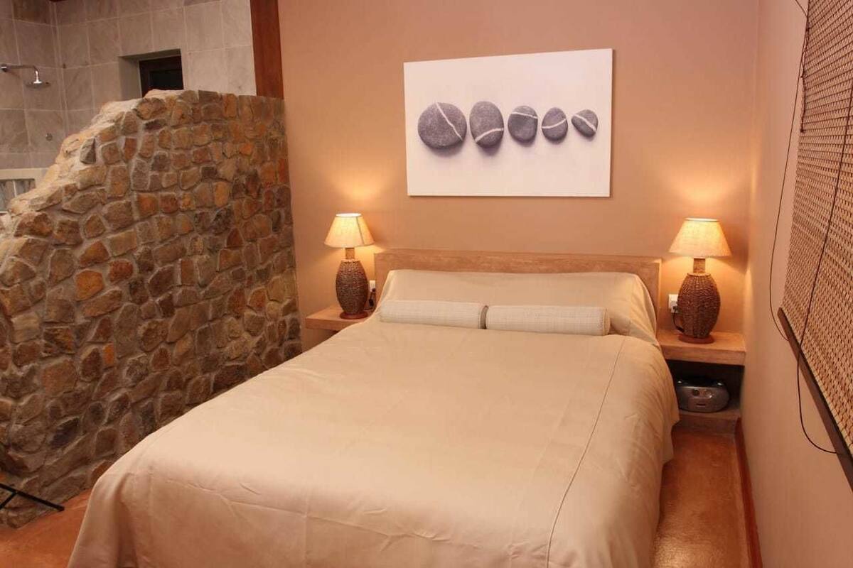 A bedroom with a stone wall, a bed with a beige bedspread, two bedside lamps, and a wall-mounted artwork of stones.