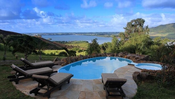 A private pool with lounge chairs, surrounded by stone tiles and natural landscaping, overlooking a lake and hills. - La Hacienda Old Grand Port Mauritius (Vieux Grand Port)