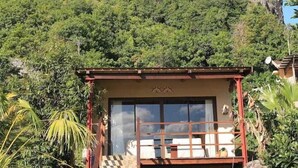 A small, rustic building with a checkered floor and a glass door, surrounded by lush greenery and a large mountain in the background. - La Hacienda Old Grand Port Mauritius (Vieux Grand Port)