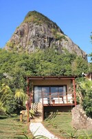 Un pequeño edificio rústico con piso a cuadros y puerta de vidrio, rodeado de exuberante follaje y una gran montaña al fondo.