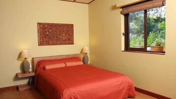Un dormitorio con cama, dos lámparas de noche, una ventana con vista a follaje y un adorno de pared.