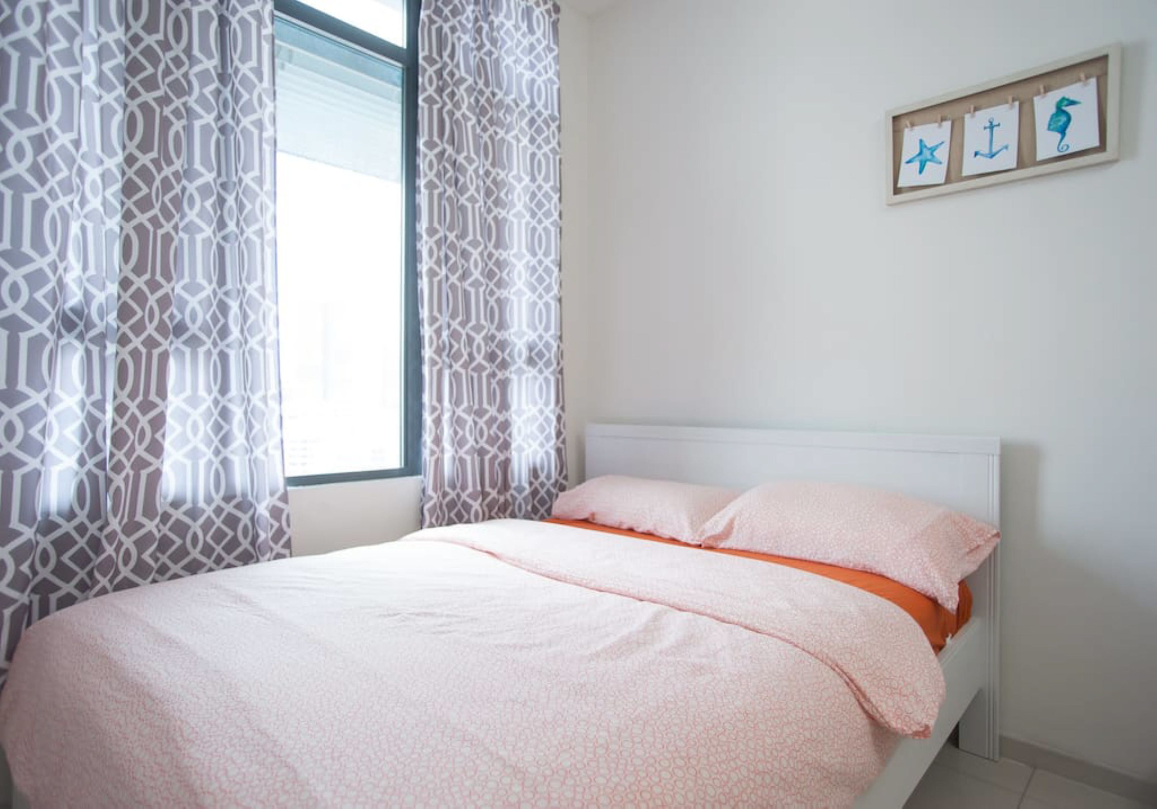 A neatly made bed with a pink and orange bedspread, a white headboard, and a window with patterned curtains.