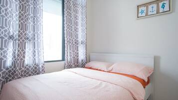 A neatly made bed with a pink and orange bedspread, a white headboard, and a window with patterned curtains.