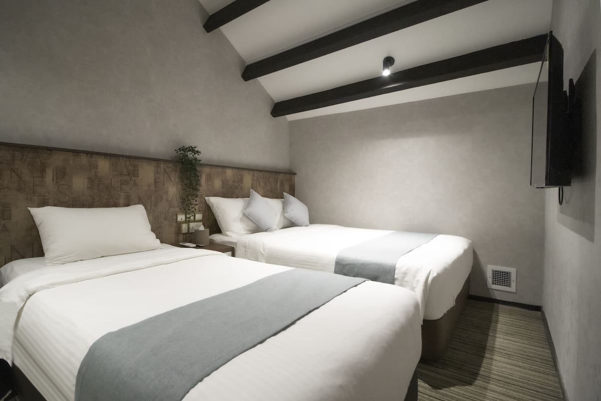 Une chambre d’hôtel avec deux lits, une tête de lit en bois, un téléviseur fixé au mur et une petite plante.