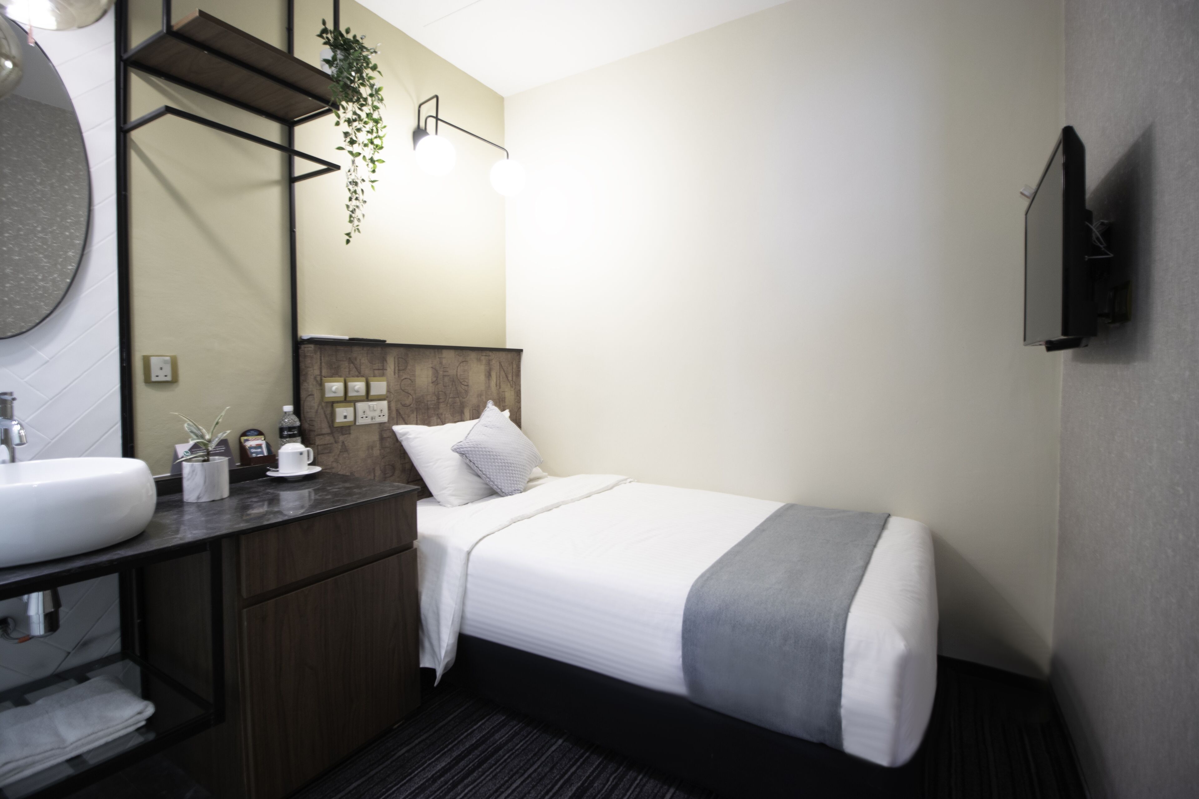 A hotel room with a large round mirror, a wooden vanity, a bed with white bedding and a gray throw, and a mounted flat-screen TV.