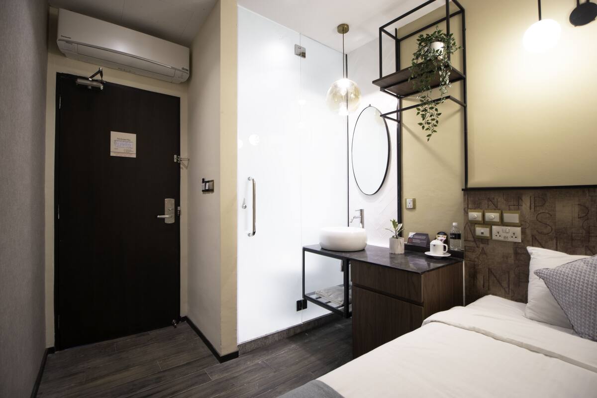 Une chambre d’hôtel moderne avec un plancher en bois foncé, un lit blanc, un petit bureau doté d’un lavabo blanc, un miroir, un luminaire suspendu et une étagère murale ornée de plantes.