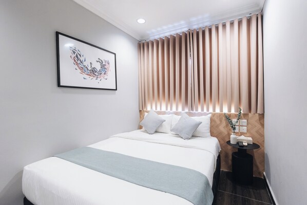 Une chambre d’hôtel moderne avec un lit soigneusement fait, une table de chevet avec une lampe et une fenêtre équipée de stores verticaux.