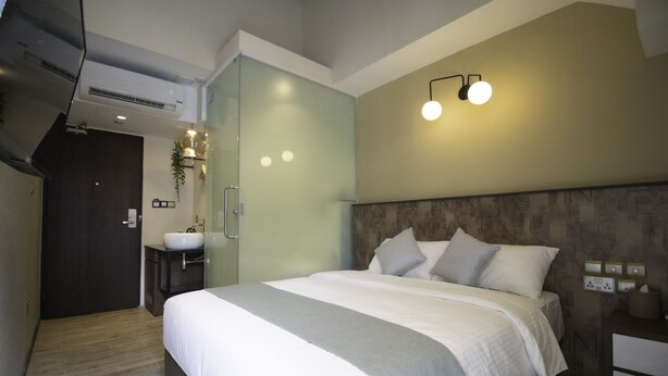 A modern hotel room with a large bed, a bedside table, a nightstand, a built-in wardrobe, a bathroom with a sink and mirror, and a wall-mounted light fixture. - Harbour Ville Hotel Hamilton - Lavender (Singapore)