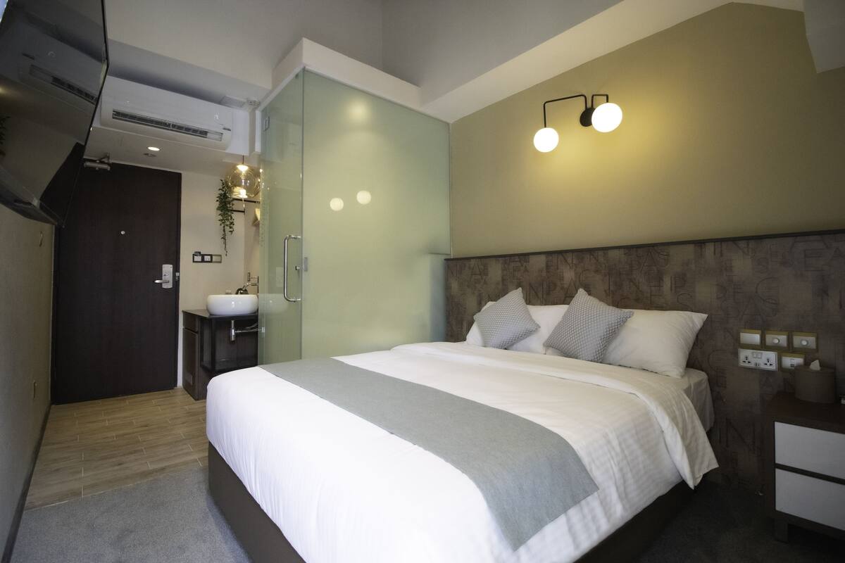 Une chambre d’hôtel moderne dotée d’un grand lit, d’une table de chevet, d’une commode, d’une armoire intégrée, d’une salle de bain avec un lavabo et un miroir, et d’un luminaire fixé au mur.