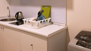 Una kitchenette compacta con un lavamanos, una tetera, una tabla de cortar y un escurreplatos.