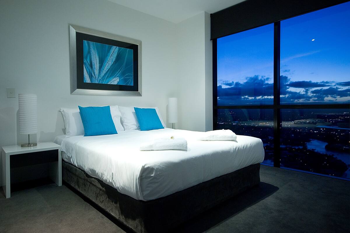 A modern hotel room with a large bed, a nightstand, a framed artwork, and a city view through large windows.