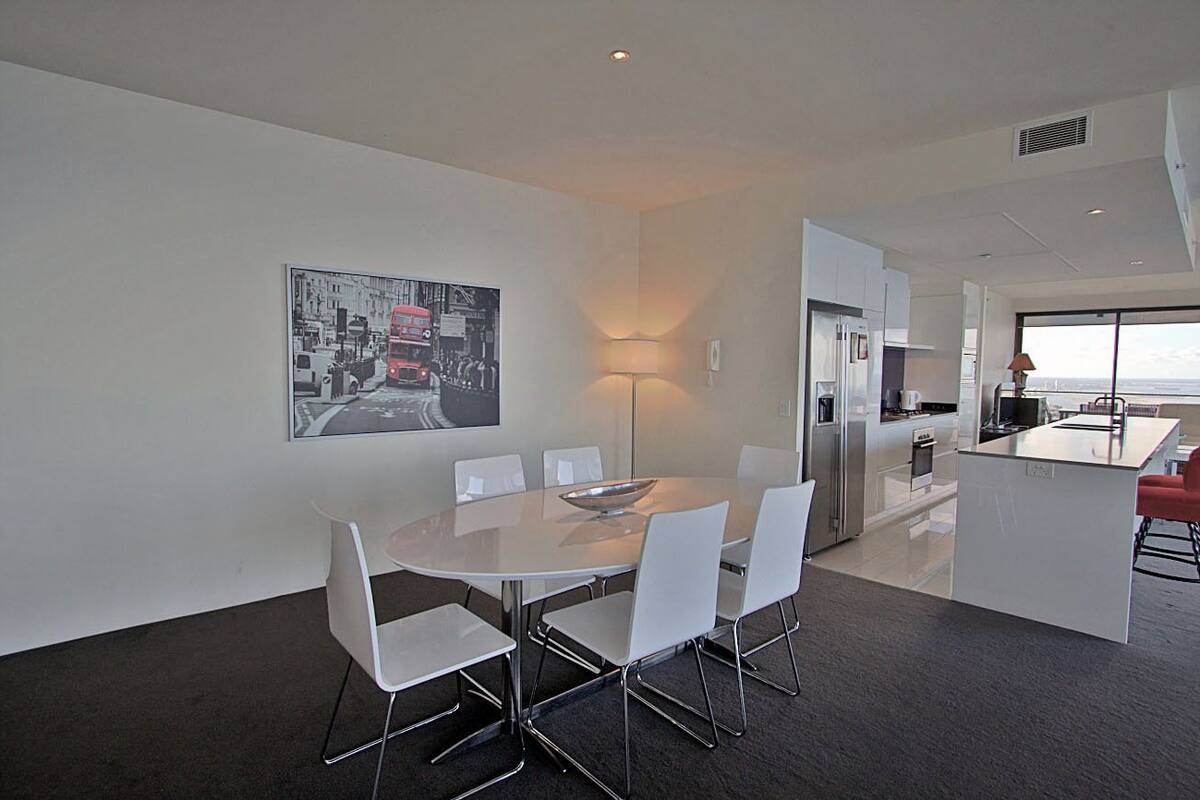 A modern kitchen with a central island, white chairs around a round table, and a cityscape painting on the wall.
