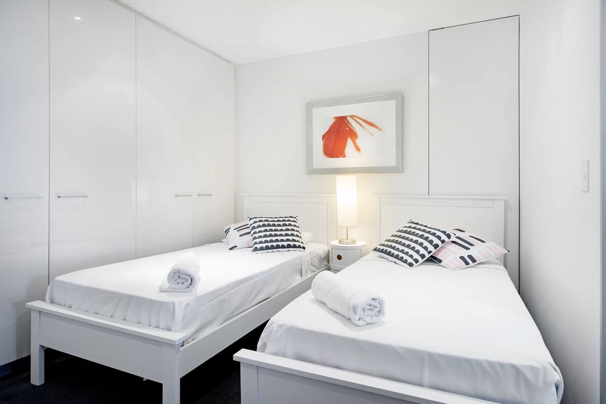 A hotel room with two single beds, white bedding, a nightstand with a lamp, and a framed picture on the wall.