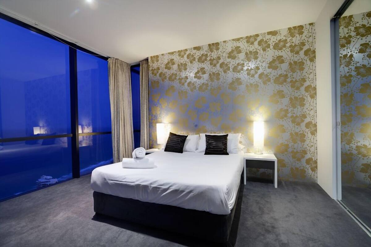 A modern hotel room with a large bed, a nightstand, and a wall with floral wallpaper.