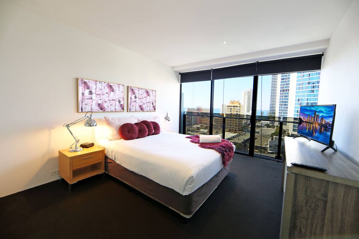 A modern bedroom with a large bed, a nightstand with a lamp, a TV, and a city view through large windows.